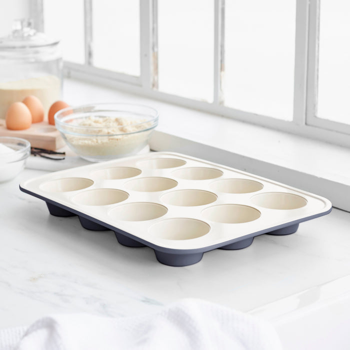 Black ceramic muffin pan with eggs and flour on a white countertop.