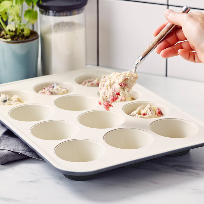 Hand scooping berry muffin batter into a black GreenLife ceramic nonstick muffin pan on a kitchen counter.