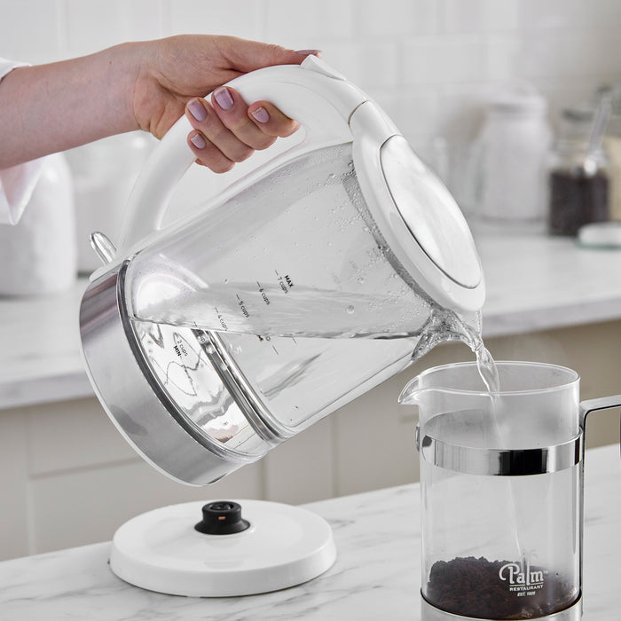 Hand pouring hot water from white glass electric kettle into a French press on a kitchen counter.
