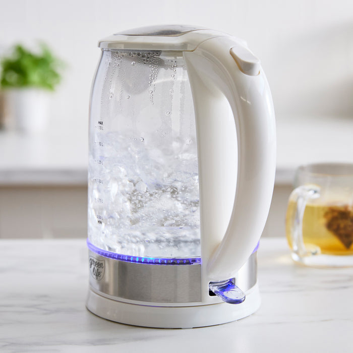 White glass electric kettle with blue LED light boiling water on a white countertop, with tea cup and potted plant nearby.