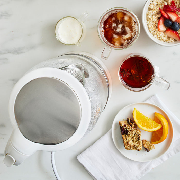 Breakfast spread with a white GreenLife glass kettle, mug of tea, iced tea, oatmeal bowl, fruit plate, and muffin.