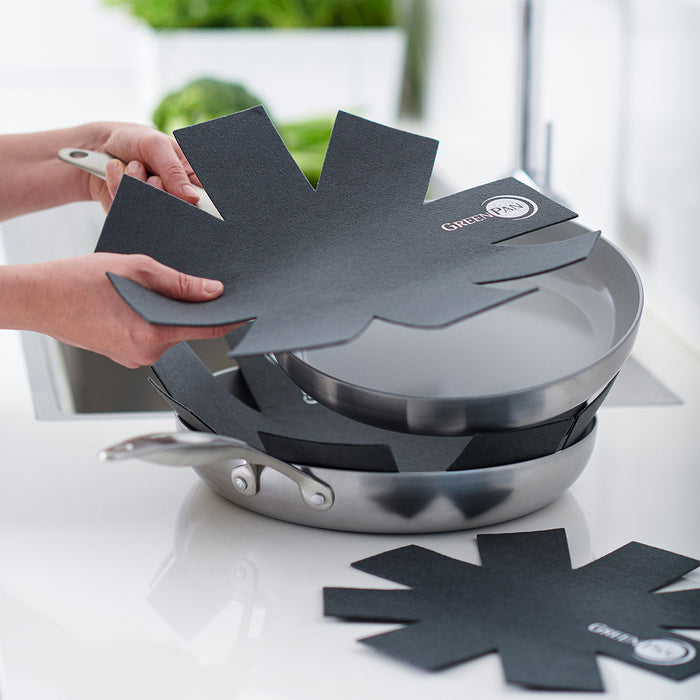 Two hands place a GreenPan 3 Pan Protector between stacked stainless steel frying pans on a kitchen countertop, helping prevent scratches. A sink and green plants appear in the background.
