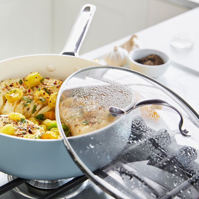 A GreenPan Glass Lid - 12", partially open, covers a pan with cooked chicken breasts topped with tomatoes, herbs, and spices. The scene includes a stainless steel handle and bowls of pepper and garlic in the background.