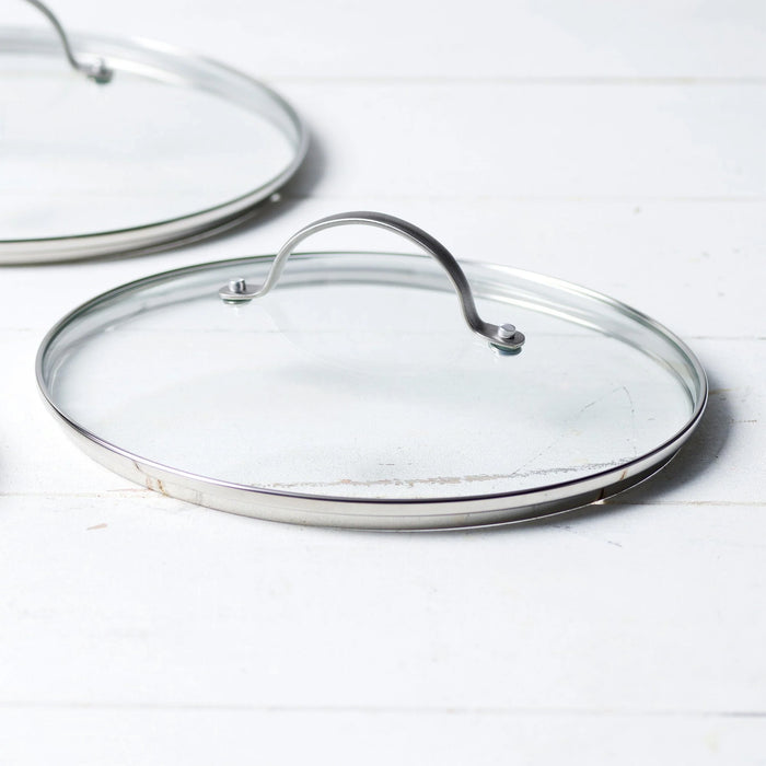 A GreenPan Glass Lid - 12" with a stainless steel handle and metal rim rests on a white wooden surface, while another similar lid appears partially in the background.