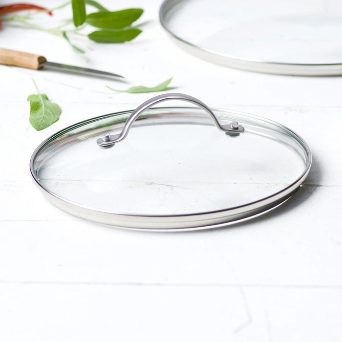 A GreenPan Glass Lid - 9" with a stainless steel handle rests on a white surface. In the background, there is a knife, green leaves, and another partially visible GreenPan lid.