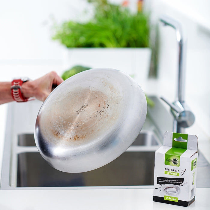 A hand washing a shiny pan over a sink with a GreenPan Melamine Sponge 2-pack box on the counter.