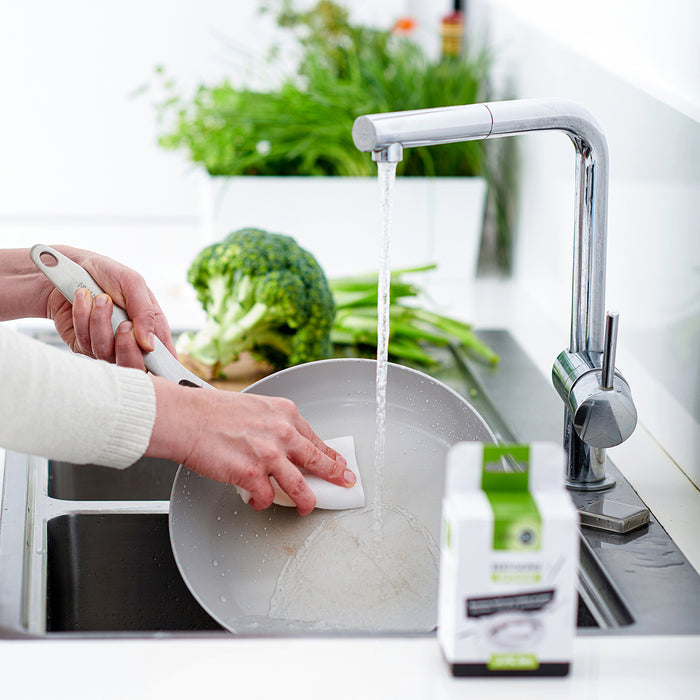 Person washing frying pan with GreenPan melamine sponge, 2-pack visible, fresh vegetables nearby on counter.