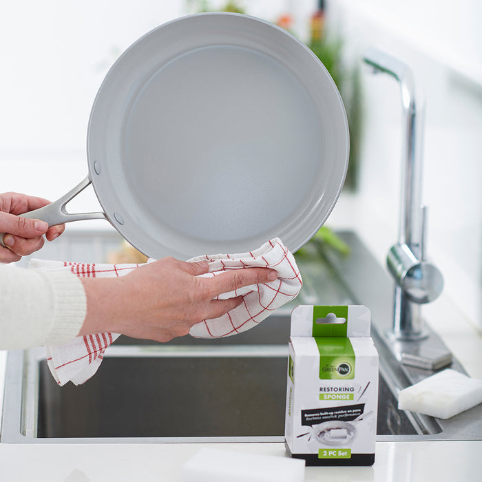 GreenPan Melamine Sponge 2-Pack box on kitchen counter next to person drying a frying pan over the sink.