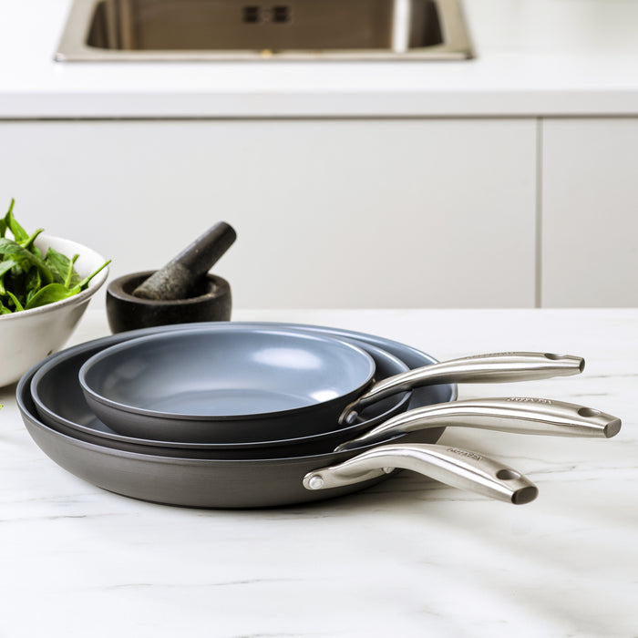 Set of three ceramic nonstick frypans in gray, stacked on a white counter beside leafy greens and a mortar with pestle.
