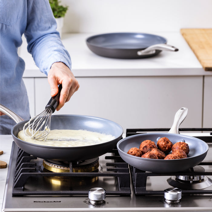 Sauce being whisked in a light green ceramic frypan, with meatballs cooking in another pan from a 3-piece set.