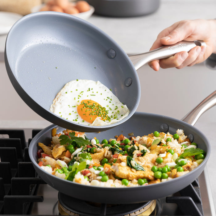 A hand slides a fried egg from a ceramic nonstick frypan onto veggie fried rice in another pan on a stove.