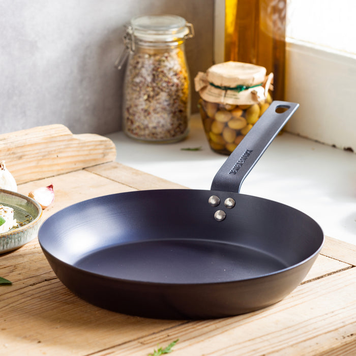 Carbon steel 10-inch frypan on a wooden counter near jars of grains and olives by a sunlit window.