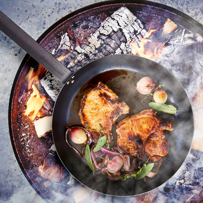 Pork chops, onions, and herbs cooking in a Merten  Storck 10-inch carbon steel frypan over an open fire.