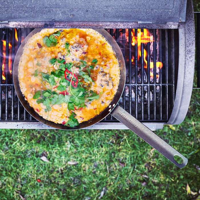 Carbon steel 10-inch frypan with stew, herbs, and chili cooking on a grill over open flame.