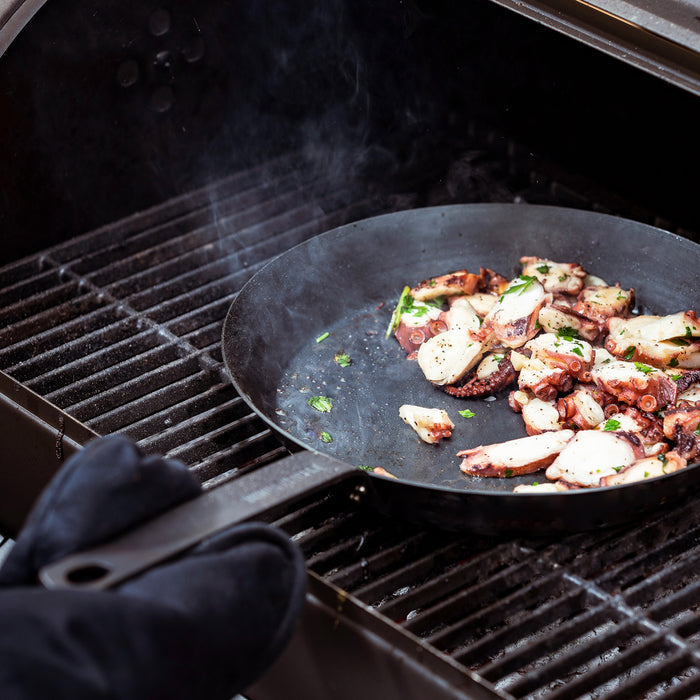 Person grilling octopus in a Merten  Storck carbon steel 10-inch fry pan, wearing black heat-resistant gloves.