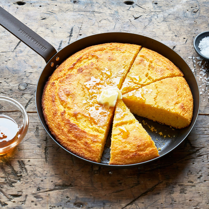Sliced golden cornbread with butter and honey in a blue carbon steel 10-inch frypan.