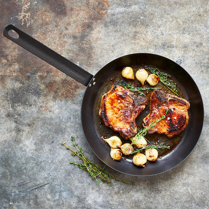 Pork chops with onions and herbs in a Merten  Storck carbon steel 10-inch frypan on a gray surface.