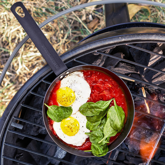 Carbon steel frypan with fried eggs, tomato sauce, and spinach cooking on a charcoal grill.