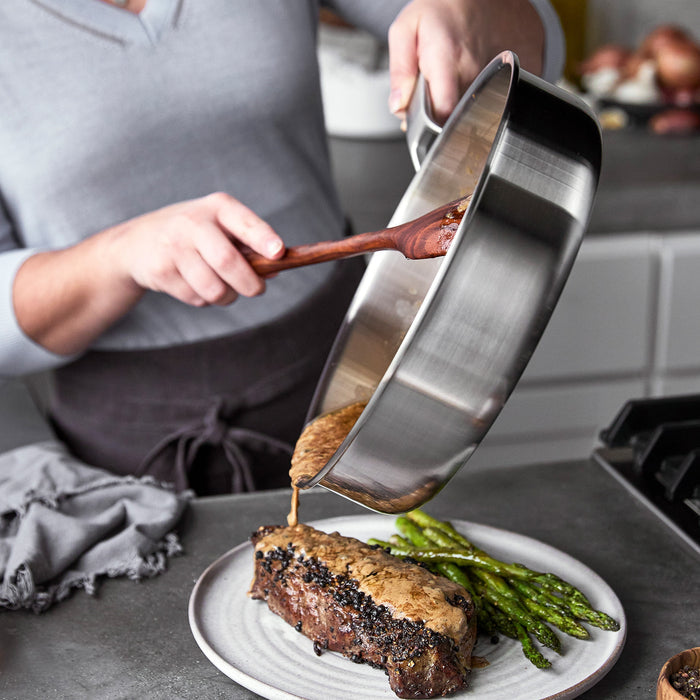 Sauce being poured in a blue Merten  Storck sauté pan onto steak and asparagus.
