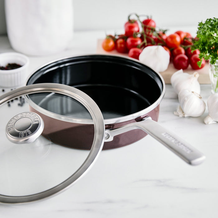 Stainless steel sauté pan with glass lid next to garlic, tomatoes, parsley, and spices on a light surface.