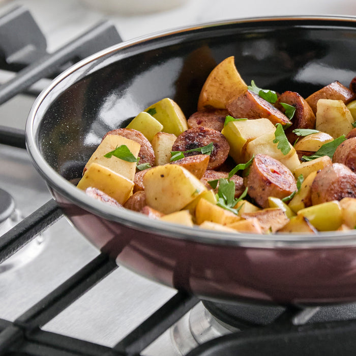 Sausage, apple slices, and herbs cooking in a brown Merten  Storck steel frypan on a stovetop.