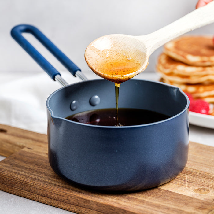 Wooden spoon drizzling syrup from a navy blue ceramic nonstick saucepan, pancakes visible in the background.