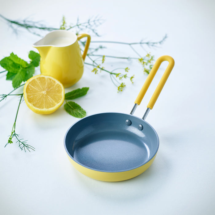 Yellow ceramic nonstick 5-inch frypan with matching handle, half lemon, white pitcher, and herbs on a light surface.