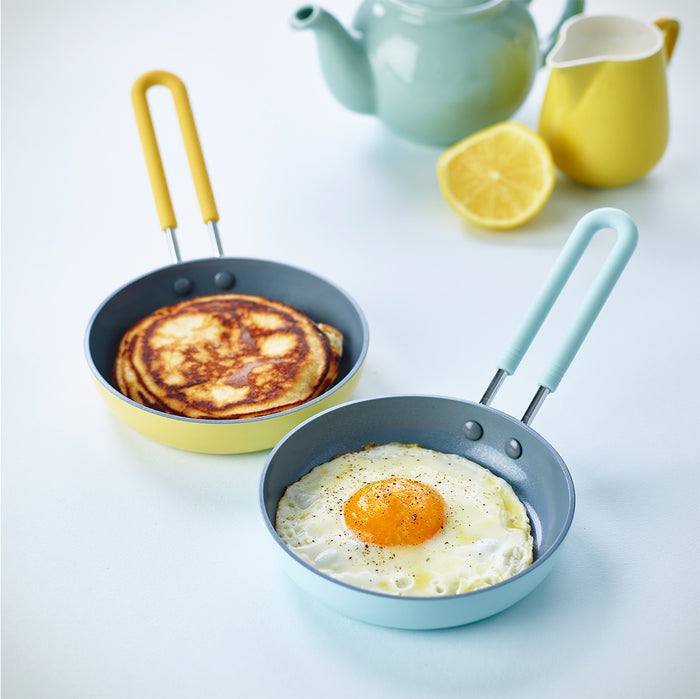 Yellow mini ceramic nonstick frypan with a fried egg and pancakes on blue surface, lemon, teapot, and creamer nearby.
