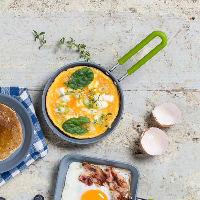 Small ceramic nonstick frying pan with a cooked omelet, spinach, herbs, eggshells, and bacon on a blue-check napkin.