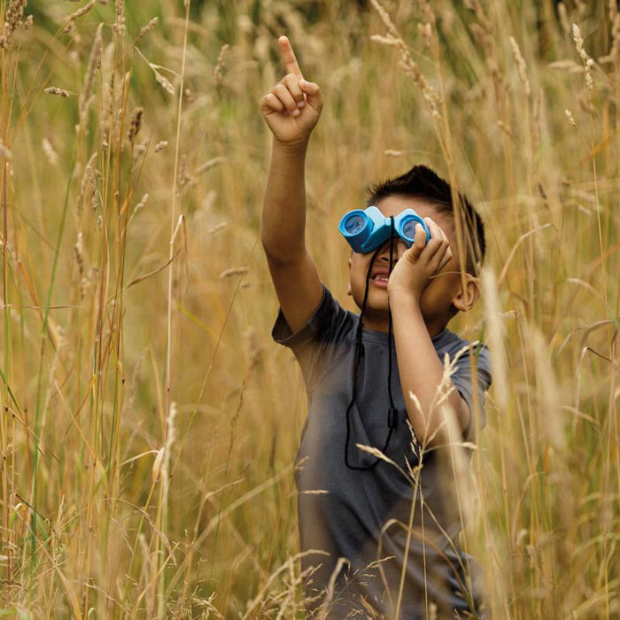 A young boy stands in tall grass, looking through the Outdoor Discovery Folding Binocular by Outdoor Discovery, pointing upward with one finger and appearing curious and engaged in outdoor exploration.