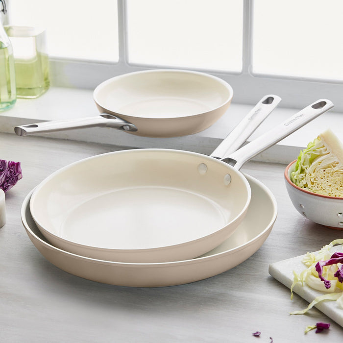 Padova Ceramic Nonstick Frypan Set in Taupe 8″, 10″, 12″ with white handles stacked on a kitchen counter by veggies and window.