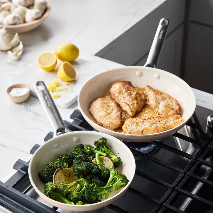 Two taupe ceramic nonstick frypans, 8″ and 10″, on a stove with chicken, broccoli, lime; lemons and mushrooms nearby.