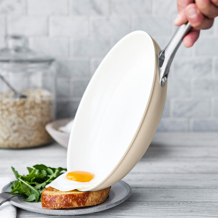 Hand holding a taupe ceramic frypan as a fried egg slides onto toast with greens.