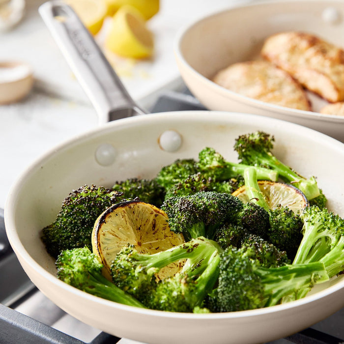 Broccoli and lemon cooking in a taupe ceramic nonstick frypan on a stove, with another pan blurred in the background.