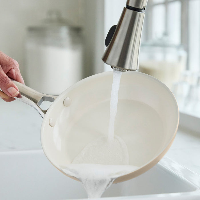 Hand washing a white ceramic frypan with soapy water in a bright kitchen sink.