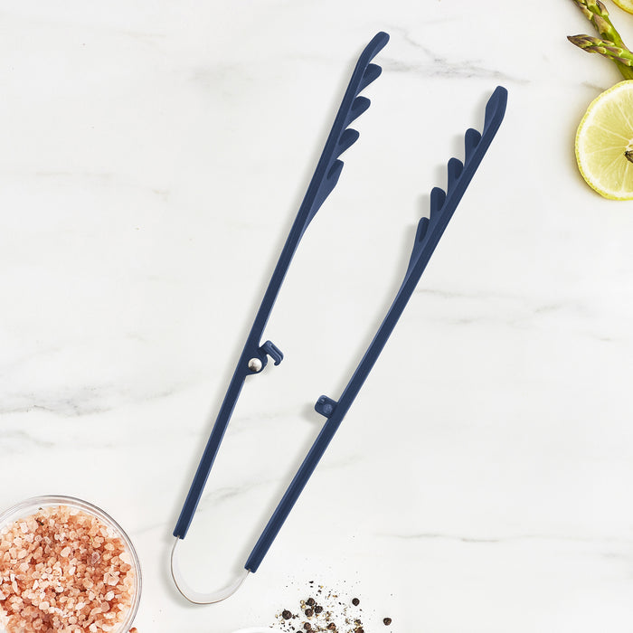 Navy silicone pasta tongs on white marble next to salt, pepper, lime wedges, and raw asparagus spears.