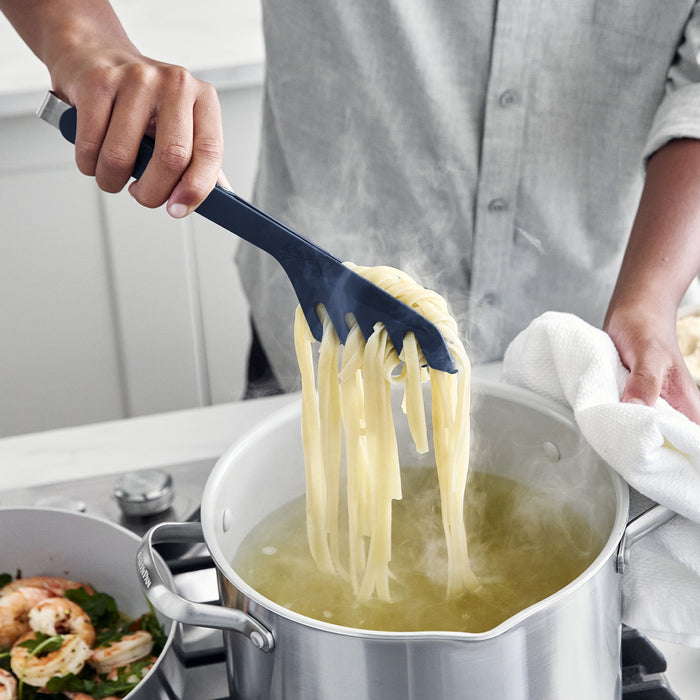 Navy platinum silicone pasta tongs lifting noodles from a steaming pot, shrimp cooking nearby on the stove.