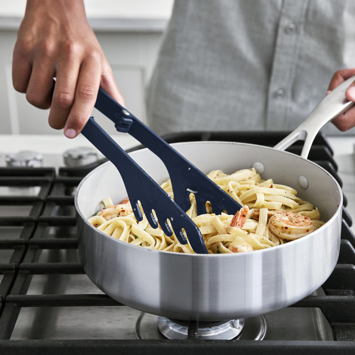 Navy platinum silicone pasta tongs mixing fettuccine and shrimp in a stainless steel pan on a stove.