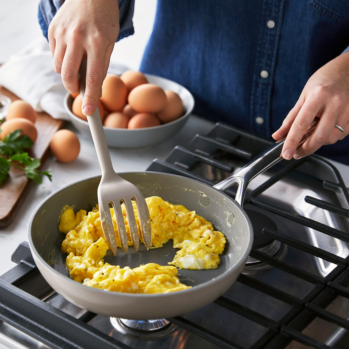 Person in a denim shirt scrambles eggs with a taupe silicone fork; eggs and parsley visible nearby.