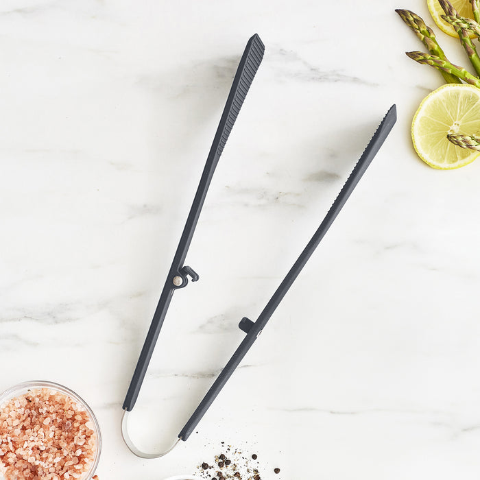 Gray silicone kitchen tongs on a white marble surface with asparagus, lemon slices, salt, and pepper nearby.