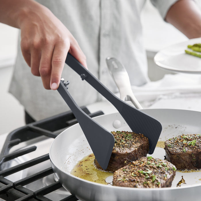 Gray platinum silicone wide tongs flipping steak pieces in a frying pan on a stovetop.