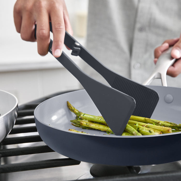 Gray silicone wide tongs holding asparagus in a nonstick skillet on a stovetop.