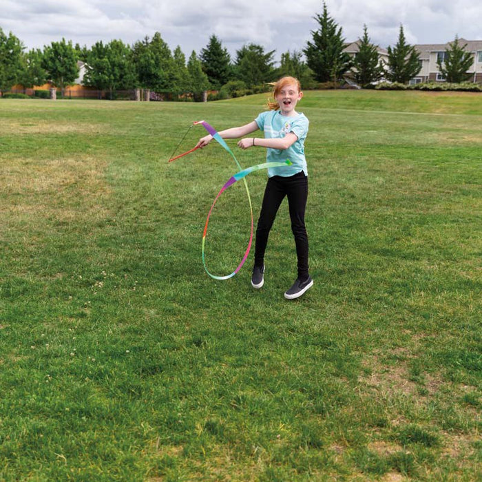 A red-haired girl in a light blue shirt and black pants plays outdoors, twirling the Playground Classics Rainbow Stunt Streamer on a grassy field with trees and houses behind her.