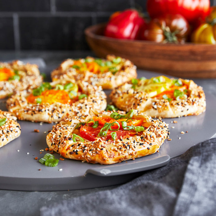 Mini bagels with tomato, green onions, and seeds on a gray round pizza baker pan; fresh tomatoes in the background.