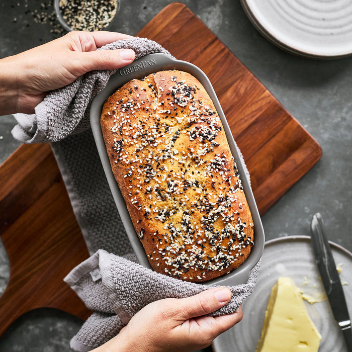 Ceramic nonstick gray 1 lb. loaf pan holding seeded bread, with butter and plates on a gray surface.