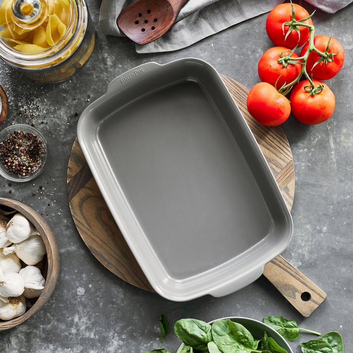 Gray ceramic 13x9 baker on a wooden board, surrounded by tomatoes, garlic, pasta, spinach, and seasonings.
