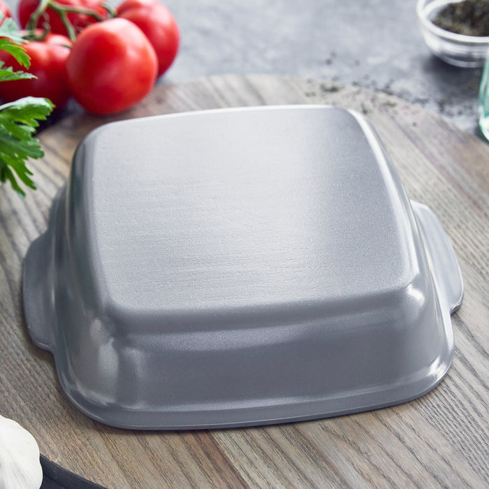 Gray ceramic square baker, upside down on wooden surface with tomatoes and herbs nearby.