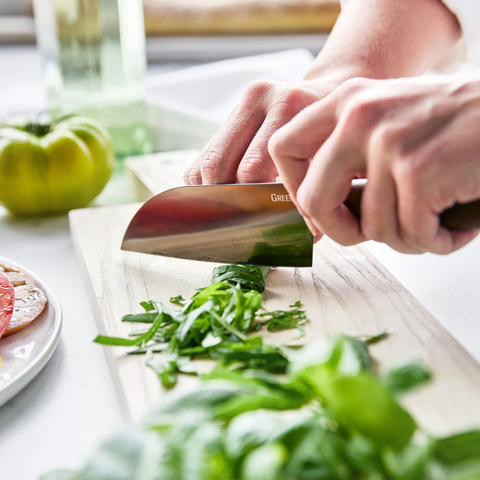 Hands chopping basil on a wooden cutting board with a silver Santoku knife from the Premiere Titanium Cutlery set.