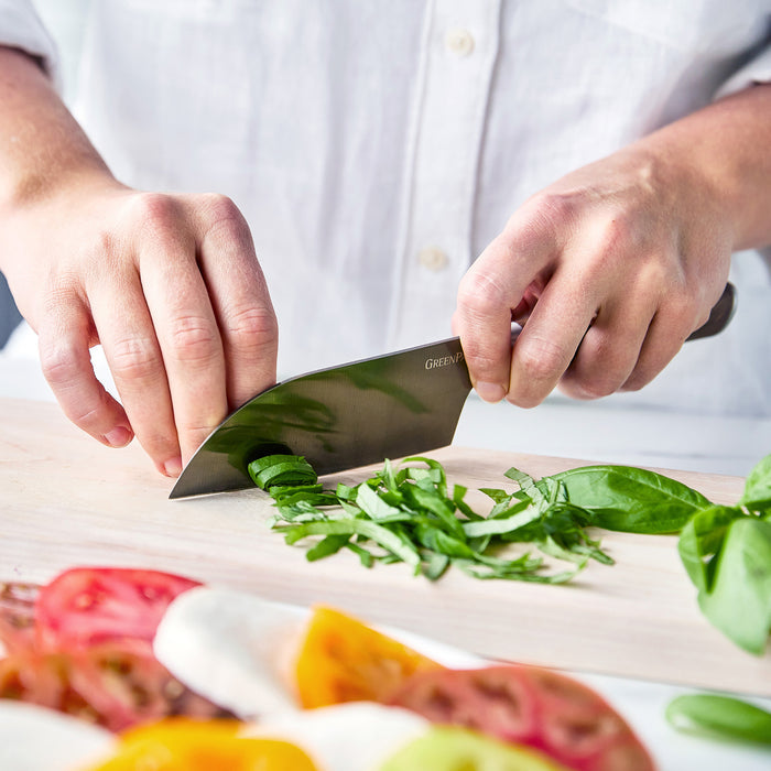 Santoku knife with a walnut handle slices green herbs on a cutting board; second knife from the set is nearby.