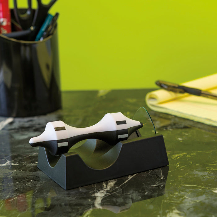 A white and black tape dispenser rests on a dark marble desk beside a pen holder, a pen, a yellow notepad, and the Project Blueprint Magnetic Levitator by Project Blueprint, with a green wall in the background.
